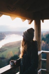 Jeune femme avec un bonnet, de dos, en train d'admirer la vue depuis le balcon d'un chalet en bois